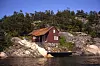 Rød hytte på svaberg ved skog og vann under klar himmel.