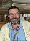Smilende mann med briller, skjegg og regnbuefarget snor innendørs.