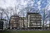 Kontor- og boligbygg bak nakne trær på en urban gate med gress foran.