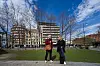 To personer står i en bypark med trær, gress og bygårder i bakgrunnen.