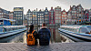 Et par sitter ved en kanal i Amsterdam med båter og fargerike hus i bakgrunnen ved solnedgang.