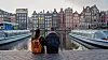 Et par sitter ved en kanal i Amsterdam med båter og fargerike hus i bakgrunnen ved solnedgang.