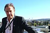 Mann i mørk dress står foran en utsikt over bygninger og åser under klar himmel.