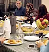 Flere personer sitter rundt et bord med serverte retter, flasker og frukt i et innendørs rom.