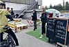 Folk står ved en markedsbod med tavleskilt, paller og parasoll på et utendørs område.