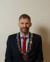 Mann i mørk dress med rød slips og seremonell kjede sitter foran en lys vegg.
