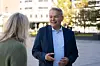 Mann i blå dress snakker med en kvinne utendørs foran en bygning.