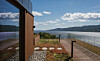 Treterrasse med glassrekkverk, steinlagt område og utsikt over innsjø og fjell under blå himmel.