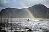 Surfer på en bølge under en regnbue med fjell og grått vann i bakgrunnen.