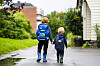 To barn med ryggsekker går hånd i hånd langs en våt vei i et boligområde.