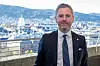 En byråd står foran et vindu med utsikt over byen og fjellene i bakgrunnen.
