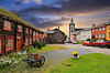 Røros kirke omgitt av historiske trehus, brostein, blomster og grønt gress under en skyfylt himmel.