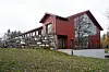 Rød trebygning med store vinduer, steinmur og løv på bakken.