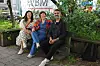 Tre personer sitter på en benk i et grønt uterom med planter og plantekasser.