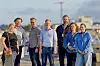 Gruppe voksne poserer ved vannet med kystlandskap og vindturbin i bakgrunnen.