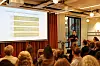 Foredragsholder ved skjerm presenterer for et sittende publikum i et konferanserom.