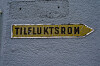 Gult skilt med teksten «TILFLUKTSROM» og pil mot høyre på en blågrå vegg.
