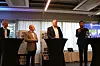Fire personer på en konferansescene med talerstol, bannere og skjerm i bakgrunnen.